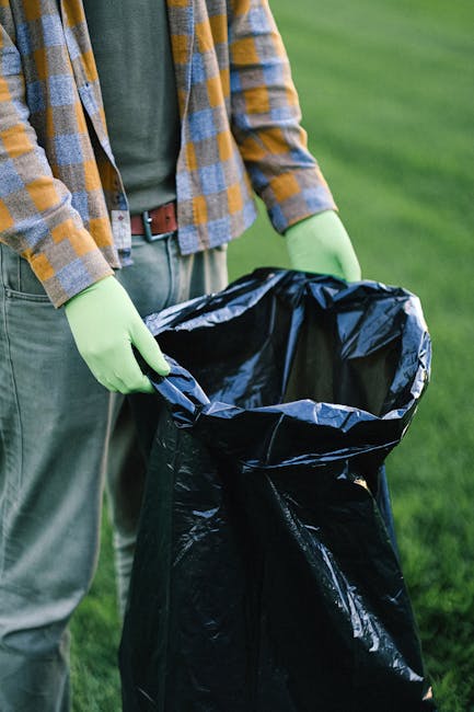 A person dressed in an orange jumpsuit standing on a concrete floor, holding two large blue plastic rubbish bags filled with waste, one in each hand. The bags are semi-transparent, revealing mixed ref