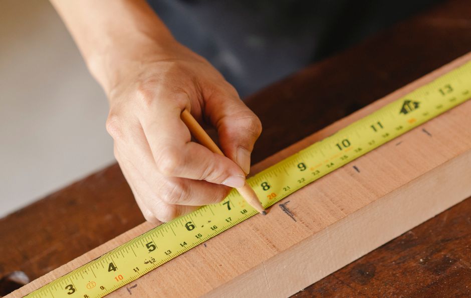 A close-up of a person's hand holding a small wooden pencil, using it to mark measurements on a light-colored wooden board placed on a darker wooden surface. The person's hand appears aged with visibl