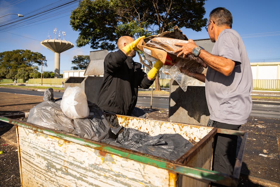 Two men are engaged in rubbish collection outdoors on a sunny day, with clear blue skies and a few trees in the background. One man, wearing a black jacket, yellow gloves, and sunglasses, is placing o