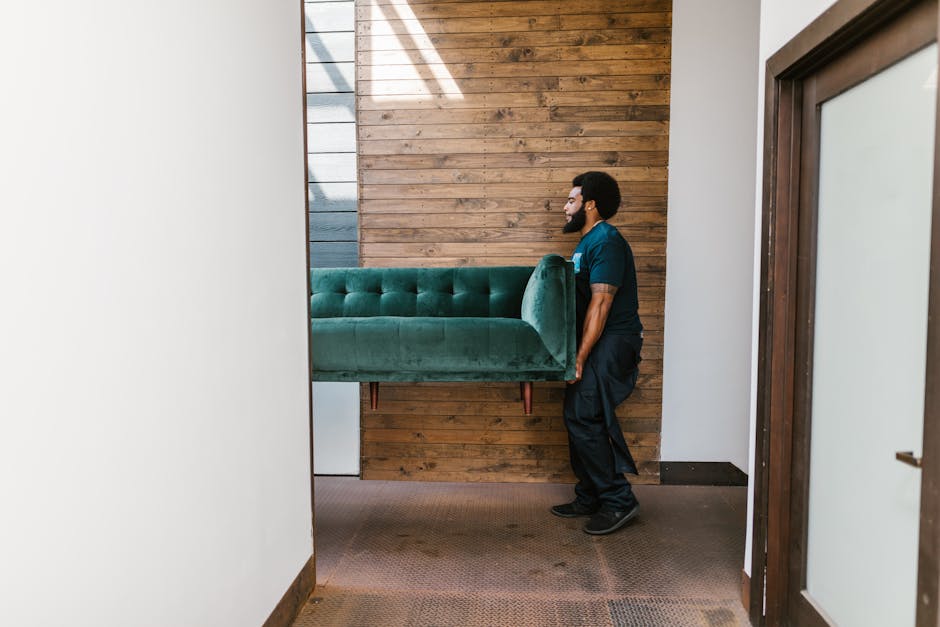 Two individuals are inside a spacious, well-lit room with white walls and large arched windows allowing natural light to fill the space. One person, with dark curly hair and a beard, is smiling while 